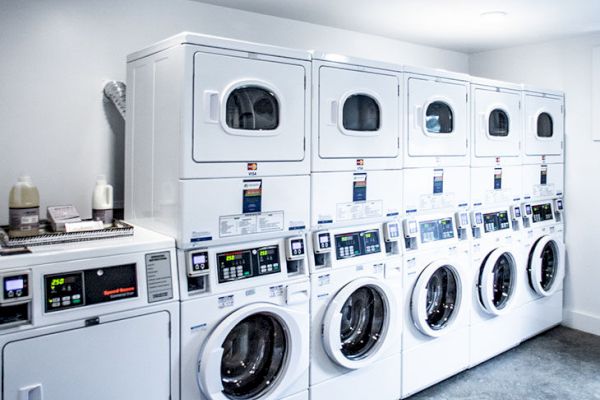 The image shows a laundry room with stacked industrial washing machines and dryers against a wall on a clean, gray floor.