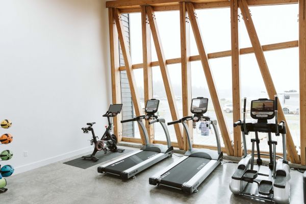 The image shows a gym with a stationary bike, two treadmills, and an elliptical machine near large windows.