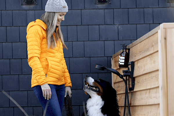 A person in an orange jacket and beanie stands beside a large dog outside near a wooden wall and outdoor shower.