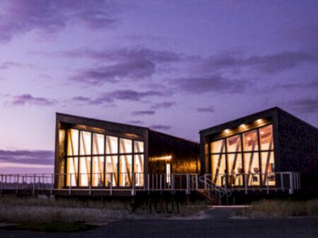 A modern building with large windows is lit up at dusk against a purple sky.