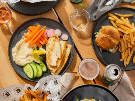 The image shows plates of food, including fries, sandwiches, veggies, salads, drinks, and sauces on a wooden table.