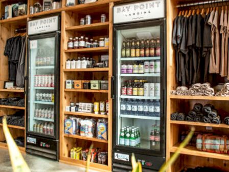 The image shows a shop interior with refrigerated displays, shelves of products, and racks of clothing. There's a plant in the foreground.