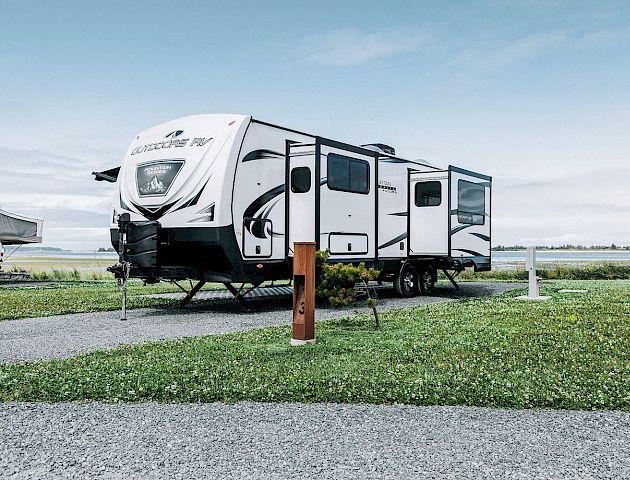 The image shows a parked travel trailer on a grassy area with a gravel path and a clear blue sky in the background.