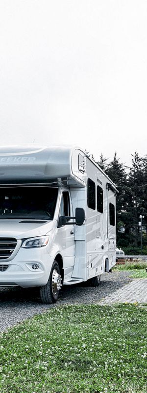 A white camper van is parked on a gravel path in a grassy campsite with trees and other RVs in the background.