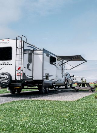 An RV is parked on a grassy lot with an awning extended. There's a picnic table nearby and another RV in the background.