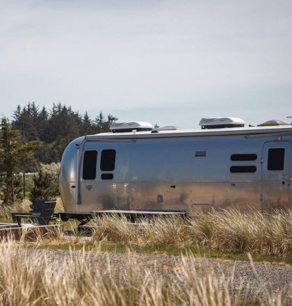 The image shows two silver Airstream trailers parked in a grassy area with trees in the background and outdoor chairs nearby.