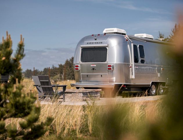 A silver Airstream trailer is parked on a grassy field, with trees and clear skies in the background, alongside outdoor chairs.