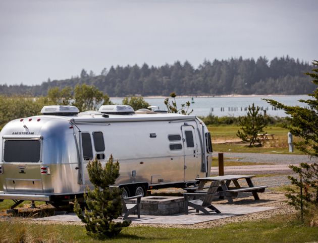 A shiny silver RV is parked near a picnic table, with a scenic lake and forest in the background, under a clear sky.