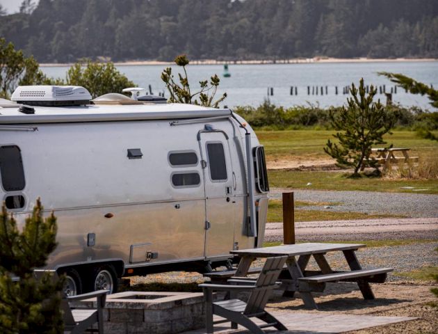 A camper trailer is parked near picnic tables by a riverside with trees and a forest in the background under a cloudy sky.