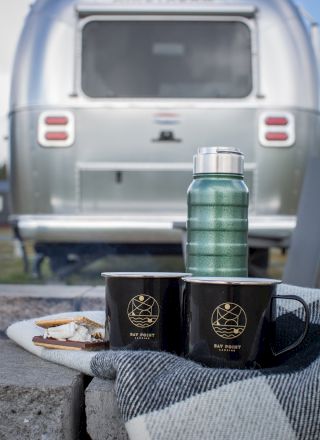 Two mugs and a thermos on a tray with snacks, set in front of a silver Airstream trailer.