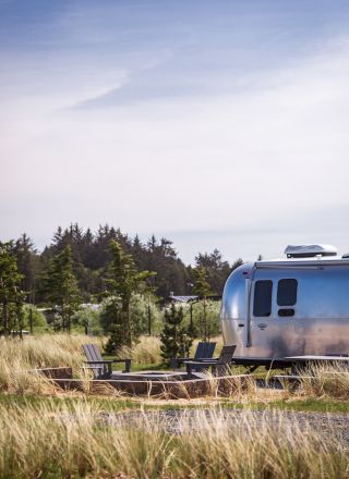 The image shows a silver Airstream trailer parked in a grassy area with trees and a clear sky in the background.