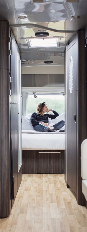 The image shows the interior of an RV with a person relaxing on a bed at the back, featuring wood accents and a small kitchen area.