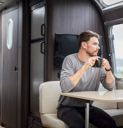 A person sits in an RV, holding a mug and looking out the window. The interior is modern and cozy, with a table and seating area.