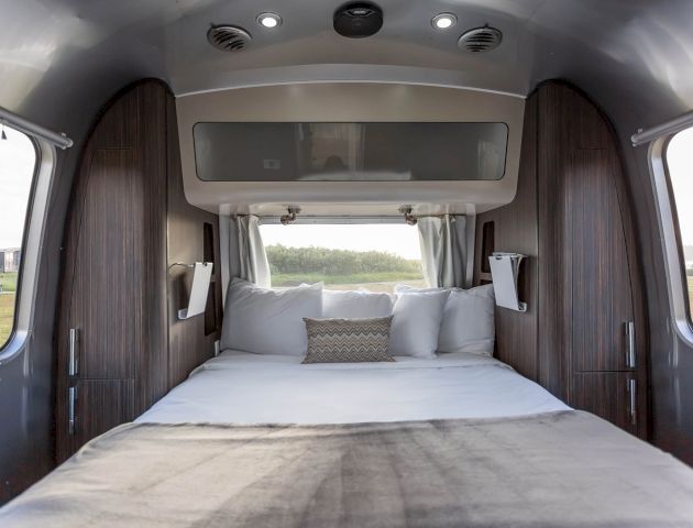 The image shows the interior of an Airstream trailer featuring a neatly made bed with multiple pillows, surrounded by wooden panels and small windows.