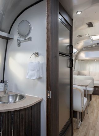 The image shows the interior of a modern Airstream trailer with a kitchenette, sink, seating area, and wooden flooring.