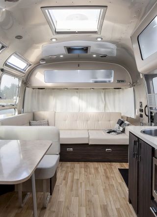 The image shows the interior of a modern travel trailer with a kitchen, dinette, and seating area. It's sleek and well-lit with ample windows.