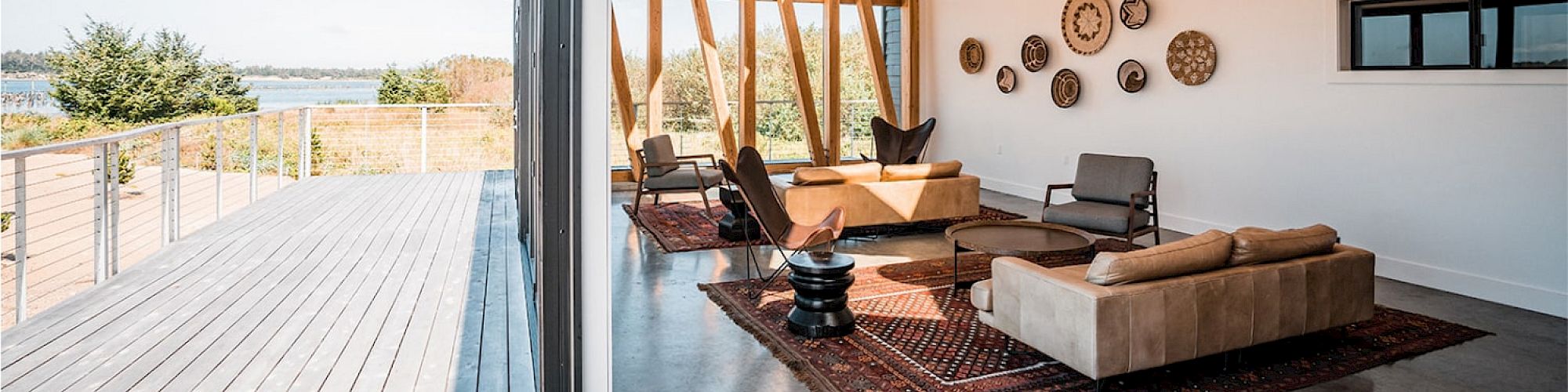 A modern living room with wooden ceilings, large windows, minimal furniture, and a deck with a view of trees and water outside.