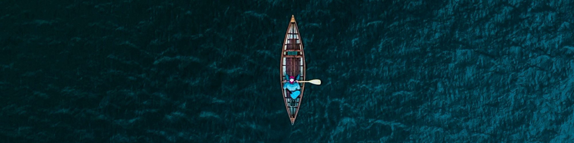 Aerial view of a lone canoeist in a small boat on a vast, dark blue body of water.