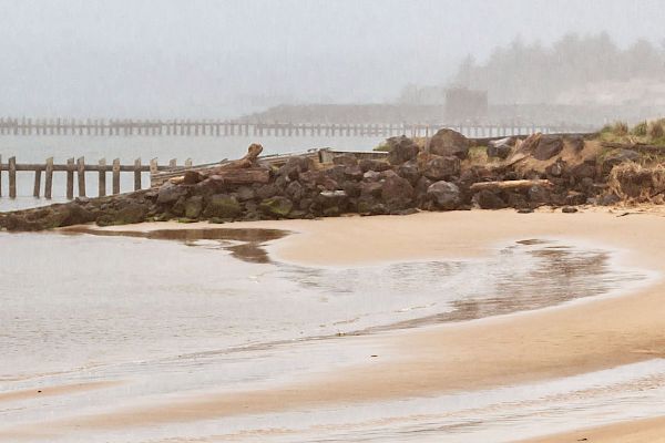 A serene beach scene with calm waves lapping at the shore, rocks, and a distant pier shrouded in a misty atmosphere.