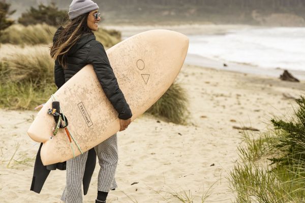 A person stands on a sandy beach holding a surfboard, wearing casual outdoor attire. The ocean and forested area are in the background.