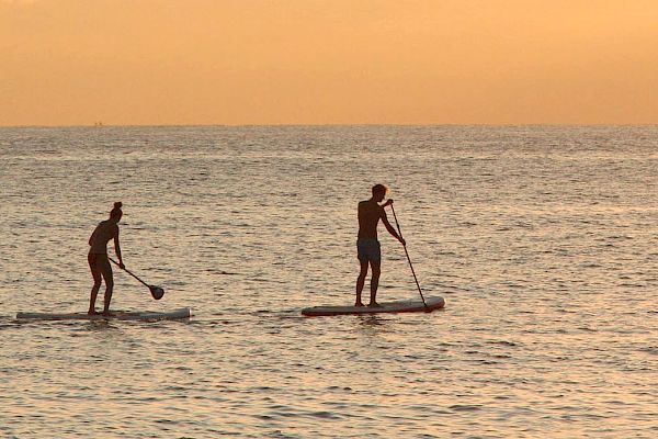 Two people are paddleboarding on the ocean at sunset with an orange sky in the background.