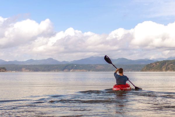 A person is kayaking on a calm lake with mountains and clouds in the background, under a bright blue sky.