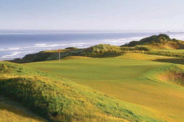 A scenic golf course with lush green hills and the ocean in the background, under a clear blue sky. The setting is serene and picturesque.