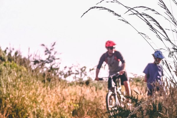 Two people are biking on a dirt path through tall grass under a clear sky, both wearing helmets.