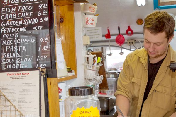 A person stands at a counter taking orders in a food place. A tip jar, menu board, and 