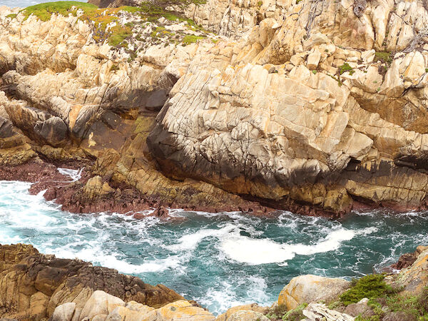 A rocky coastline with waves crashing against the shore and some greenery around. The background shows distant hills or cliffs.