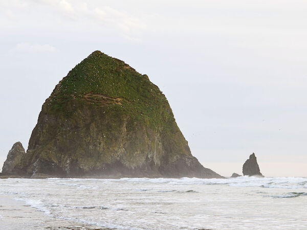 A large rock formation on a beach, with gentle ocean waves and a reflection on the wet sand.