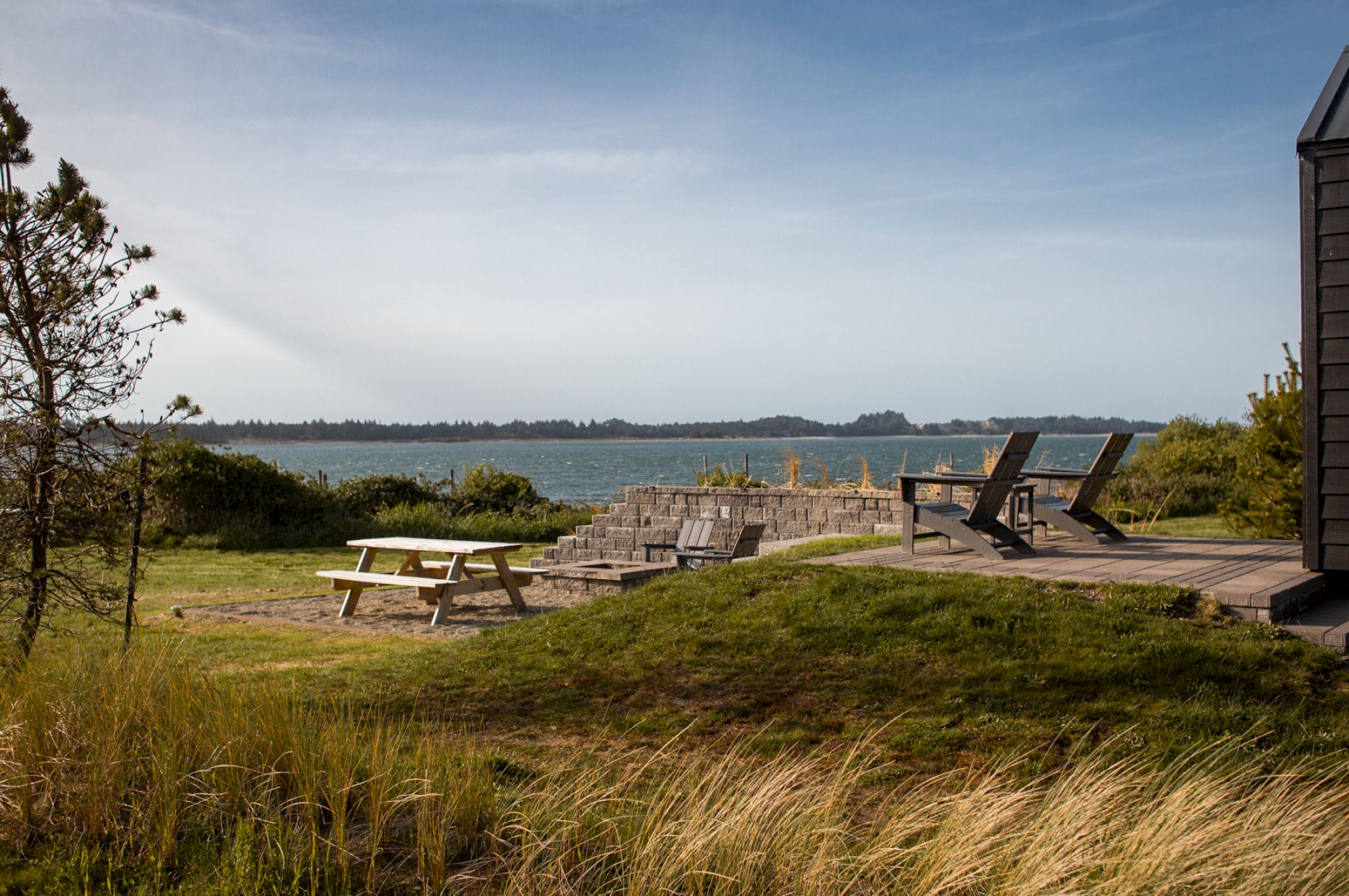 A scenic view with a picnic table, chairs on a deck, and grassy area overlooking a body of water under a clear blue sky.