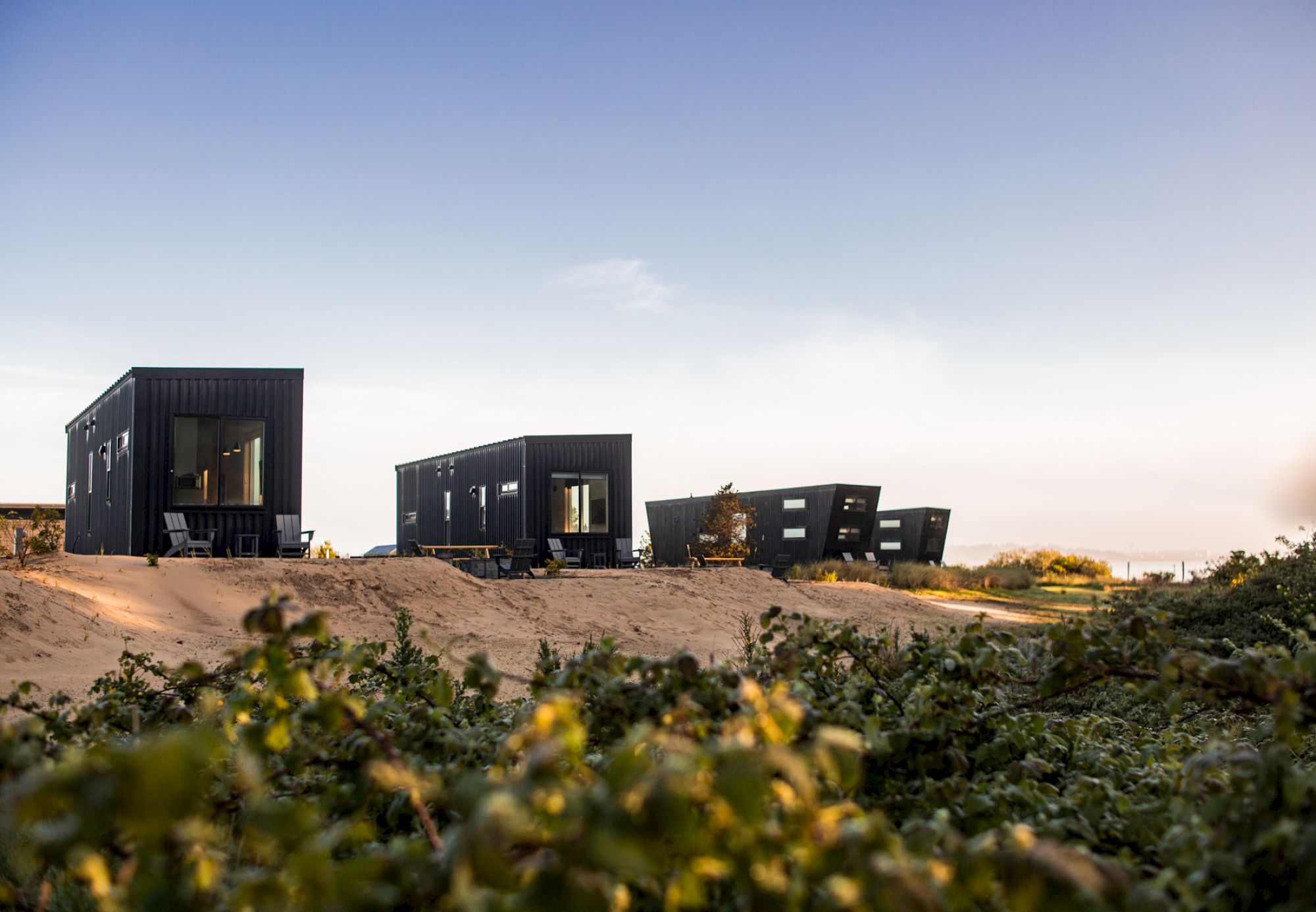 The image shows modern, black rectangular cabins in a sandy, grassy landscape under a blue sky, with a serene and minimalistic atmosphere.