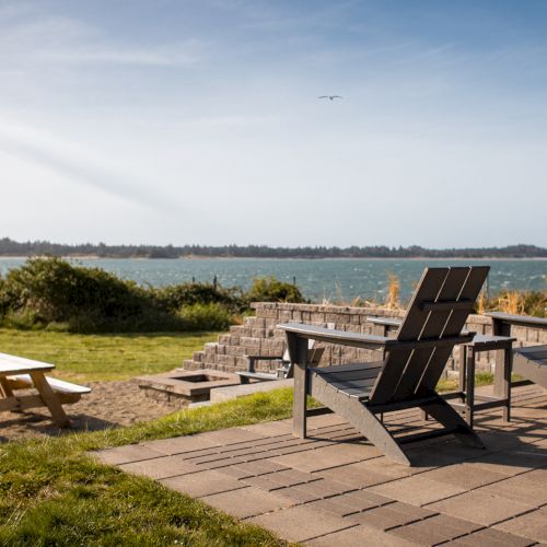 A scenic patio view with Adirondack chairs, a picnic table, and a beautiful lake in the background under a clear blue sky.