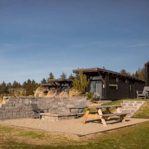 A wooden cabin with outdoor seating is set in a grassy area, surrounded by trees. A picnic table is nearby on a stone platform.