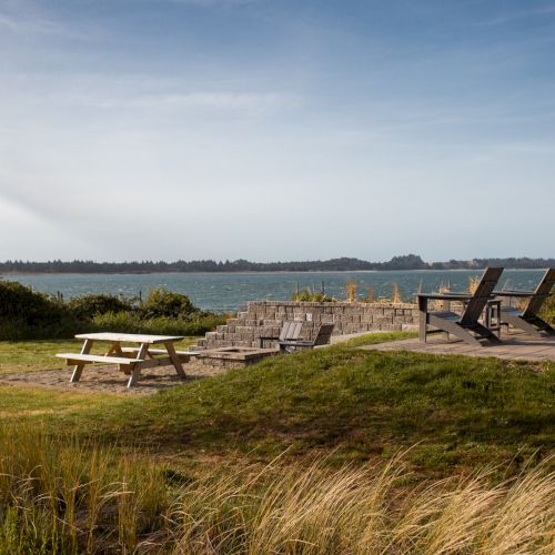 The image shows a scenic outdoor area with a picnic table, lounge chairs, and a view of the water on a sunny day.