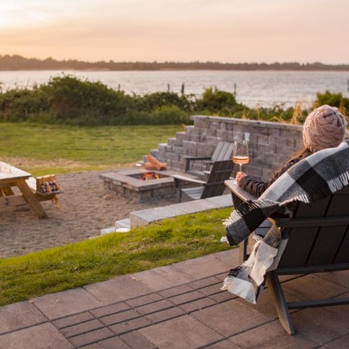 A person sits in a chair wrapped in a blanket near a fire pit, overlooking a scenic outdoor area with a picnic table at sunset.