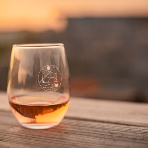 A stemless wine glass with a logo on it, partially filled with a rosé or amber liquid, sits on a wooden surface at sunset.