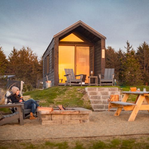 A cozy cabin with large windows is surrounded by chairs, a picnic table, and a fire pit, set against a backdrop of trees and a clear sky.