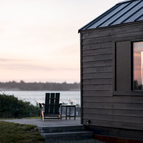 A wooden cabin with a metal roof overlooks a serene body of water at sunset, with chairs and a table on the patio outside.