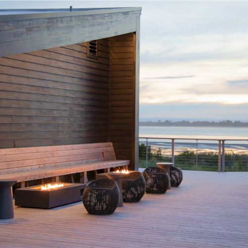 An outdoor terrace with a wooden bench, fire pits, and scenic views of the water in the background, set during dusk.