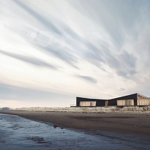 A modern building stands near a beach with a dramatic sky overhead, featuring sweeping clouds and a calm shoreline in the foreground.