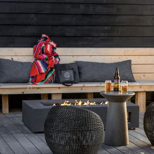 A cozy outdoor seating area with a fire pit, woven furniture, and blankets. A small table holds drinks, adding a relaxing touch.