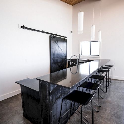 A modern kitchen with a black countertop island, bar stools, pendant lights, and wooden ceiling beams is shown in the image.