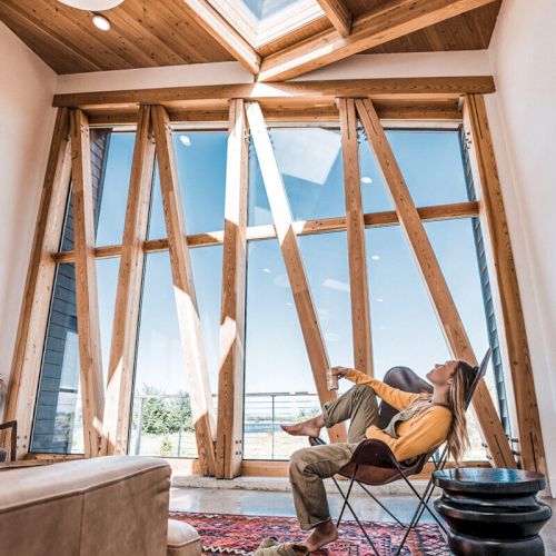 A cozy room with wooden beams, large windows, a person relaxing in a chair, rug, and a sky view through a skylight completes the scene.