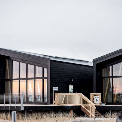 A modern building with large windows and a wooden deck, surrounded by tall grass, with a cloudy sky in the background.