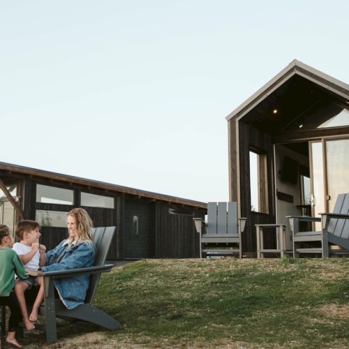 A family sits together outside on wooden chairs, beside modern cabins in a rural area. The setting is peaceful and rustic.