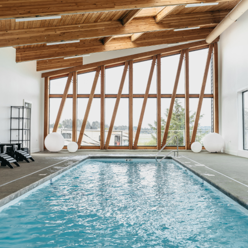 An indoor swimming pool with a modern wood beam ceiling, large windows, and seating on the side.