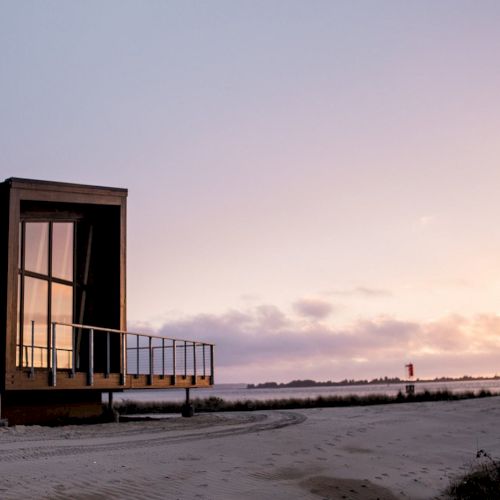 A modern building with large windows sits on a sandy beach at sunset, with a vast sky and distant horizon completing the serene backdrop.