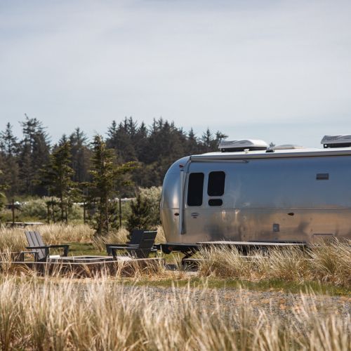 Two silver trailers are parked near a grassy area with chairs and a fire pit, set against a backdrop of trees under a cloudy sky.